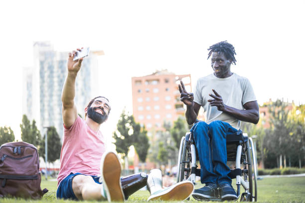 Two young adult friends taking selfie, African man on wheelchair and Hispanic man with artificial limb having a video call with friends at city park, people with disabilities lifestyle